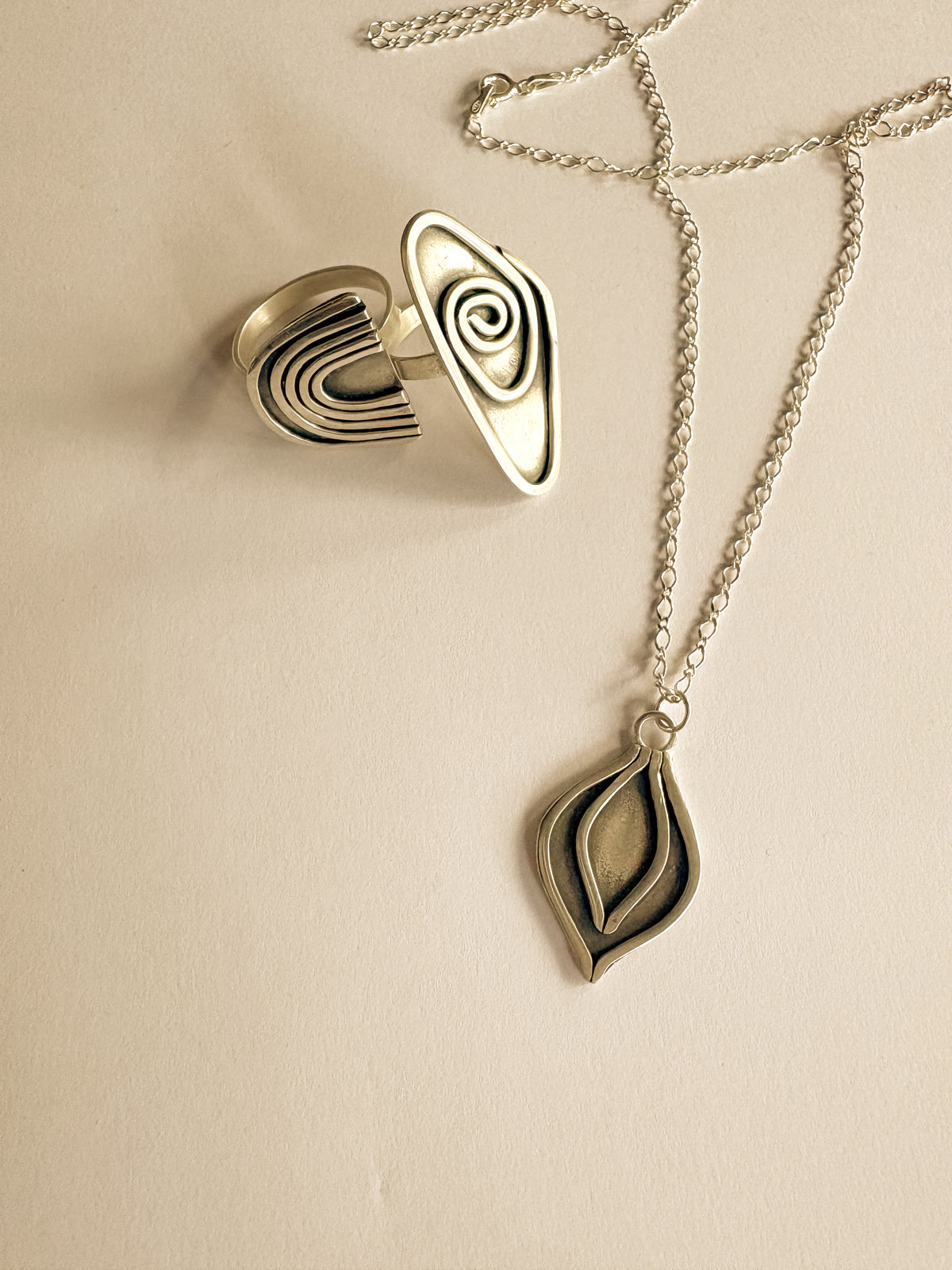 A silver-toned jewelry set featuring two rings and a necklace, both with patina  layered silver design, displayed on a warm beige background.
