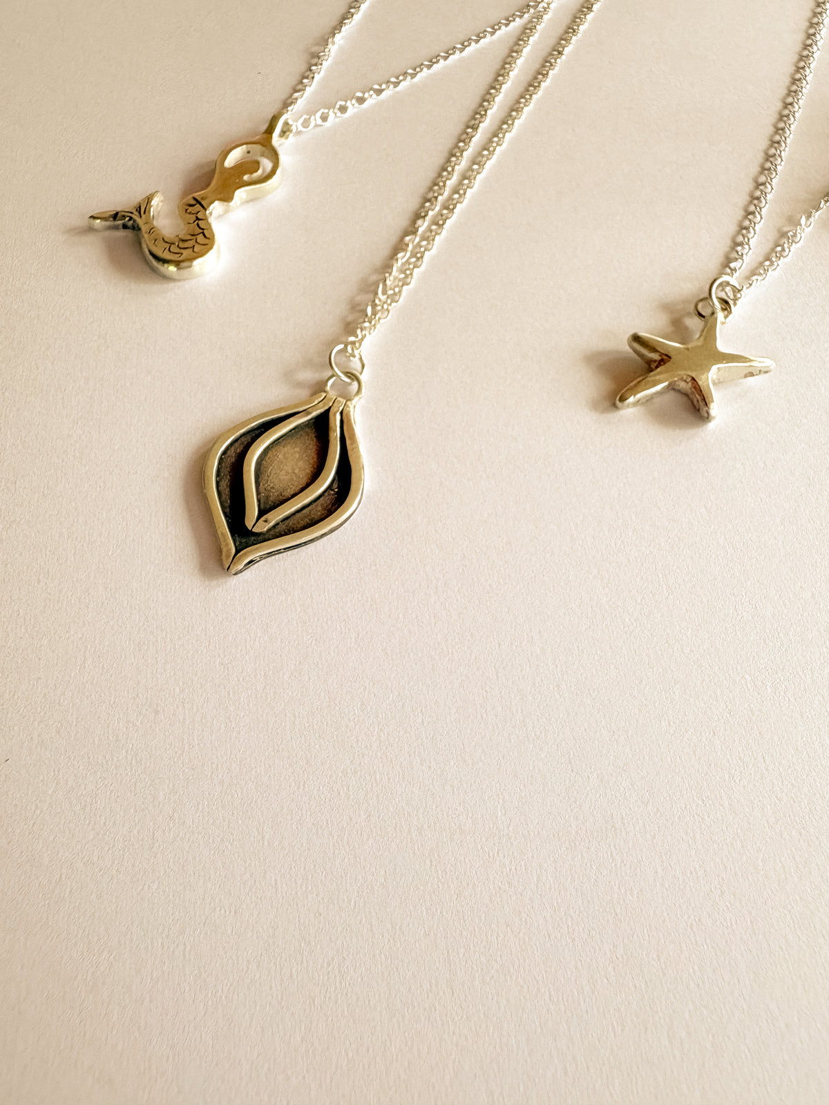 Three silver chain necklaces with unique pendants, including a mermaid, a lotus petal and a sea star, arranged on a light beige background.